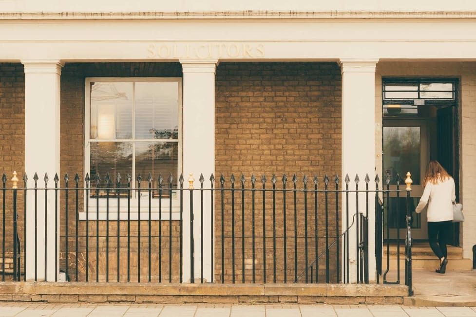 Brick building facade with black iron fence, white pillars, window, and a woman entering a doorway.
