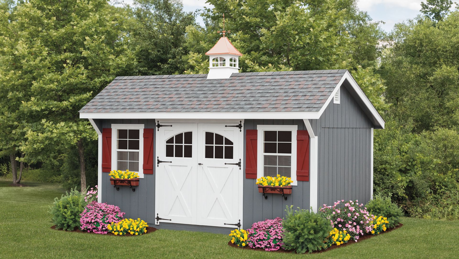 Gray shed with white doors, red shutters, and a cupola; set in a garden with flowers.