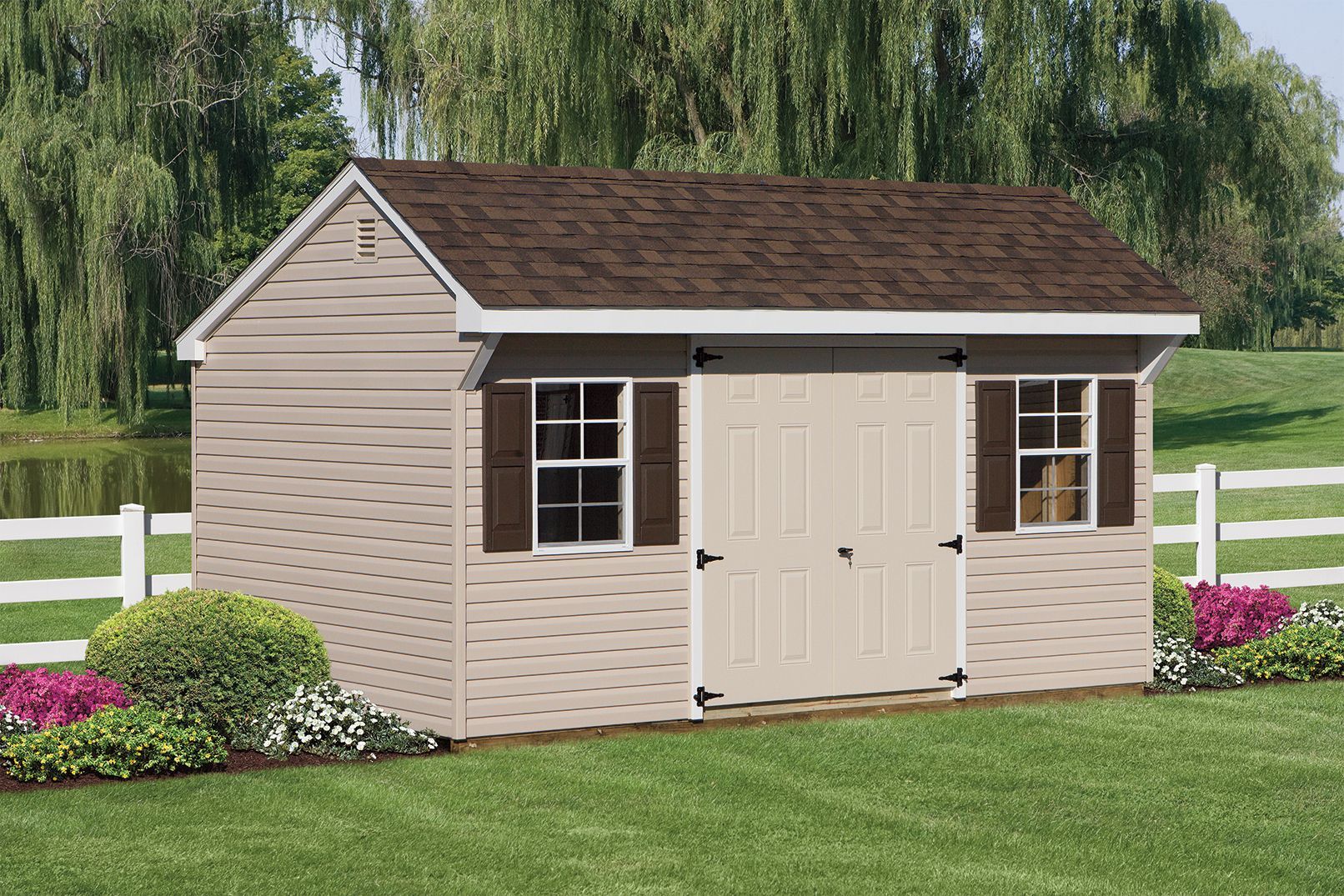 Tan shed with brown roof, white trim, two windows, and double doors, set in a yard with a white fence.