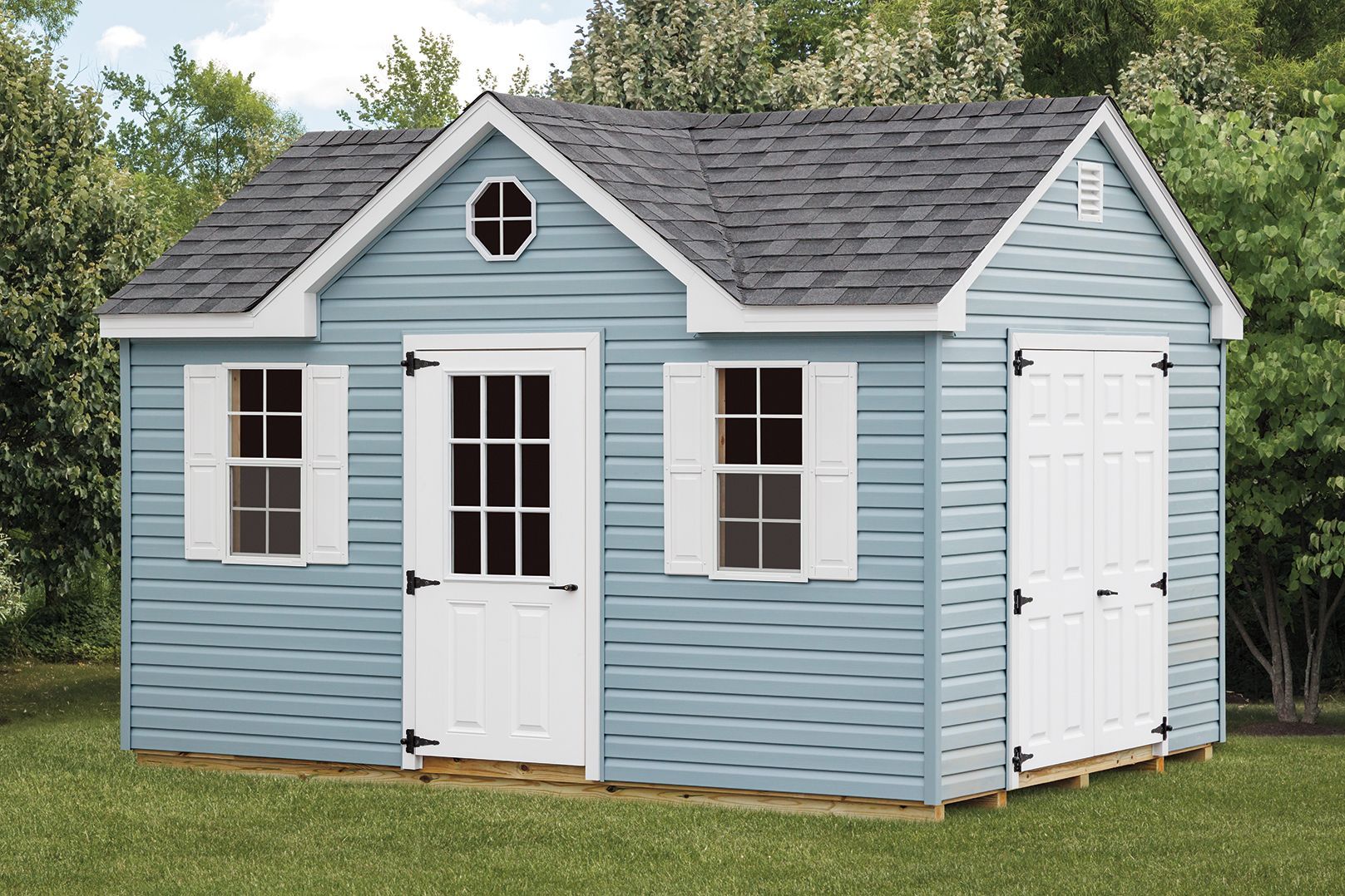 Blue shed with white trim, door, and shutters. Gray roof, small oval window in gable, set on grass.