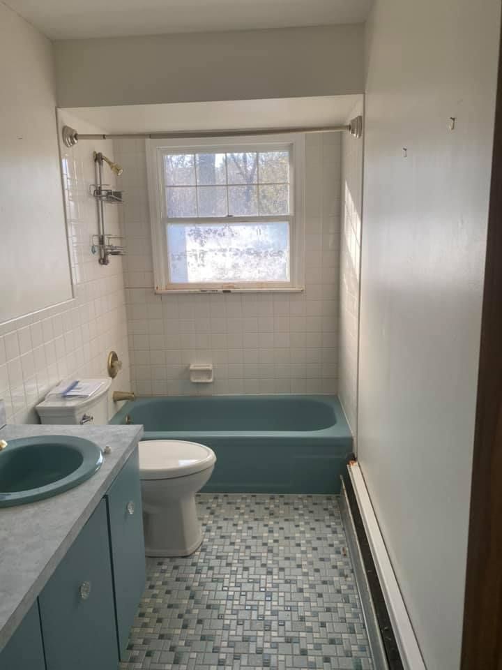 Small bathroom with teal sink, toilet, and bathtub, white tile walls, and a window above the tub.
