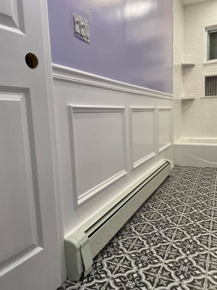 Hallway with white wainscoting, pale lavender wall, patterned carpet, and baseboard heater near a doorway