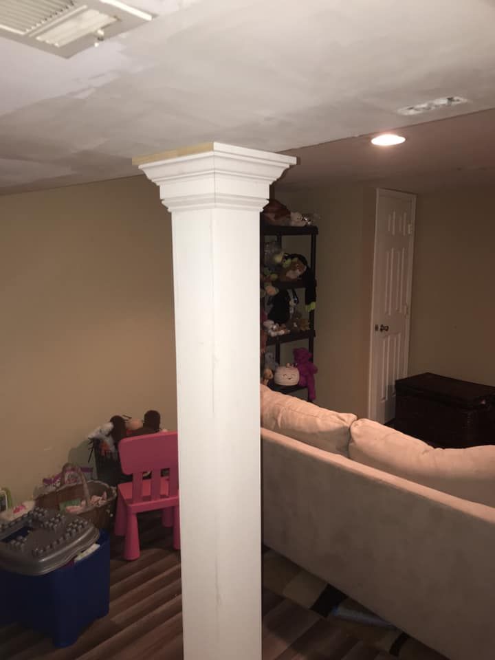 Finished basement with a beige couch, white column, storage bins, and a small pink chair under low ceiling lights