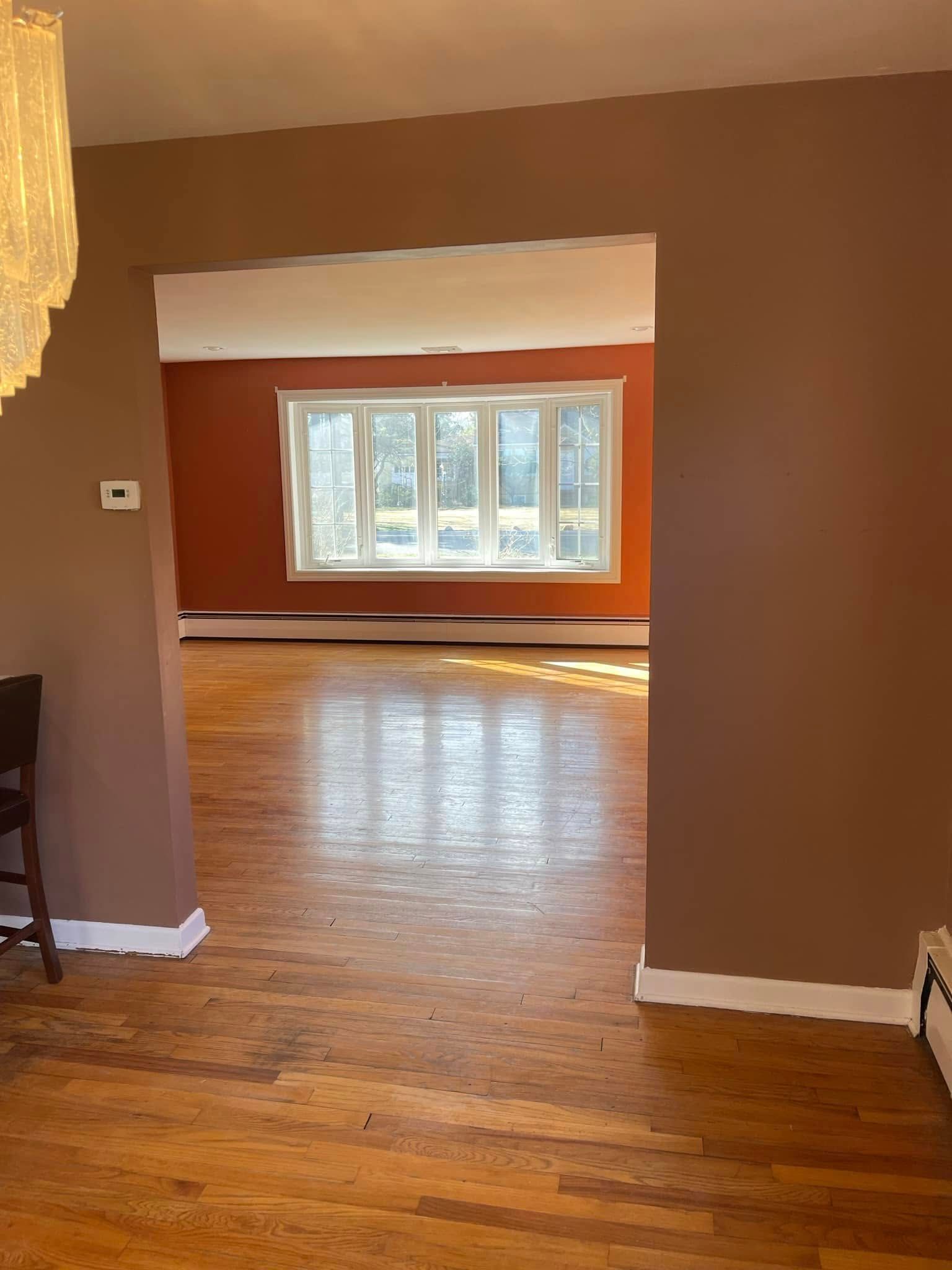 Empty living room with hardwood floors, brown walls, and a large window opening into a sunlit room