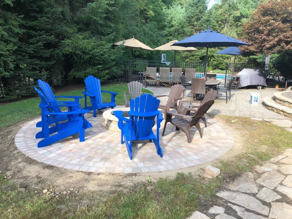 Blue Adirondack chairs around a fire pit on a stone patio in a wooded backyard.
