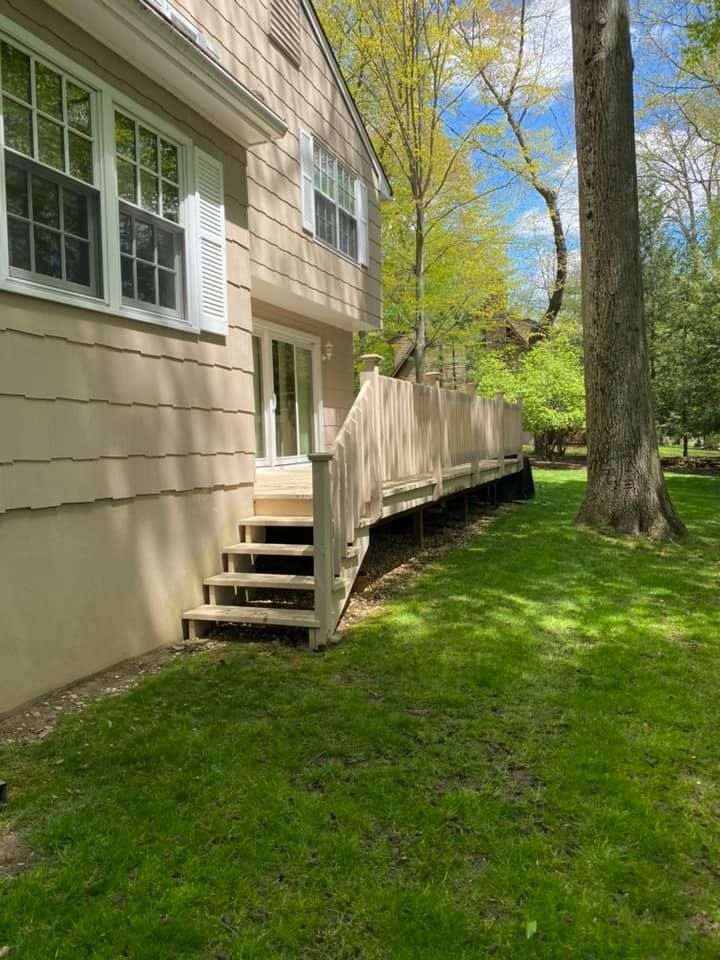Apartment exterior with tan siding, small stairs, and a wooden ramp beside a grassy yard with trees.