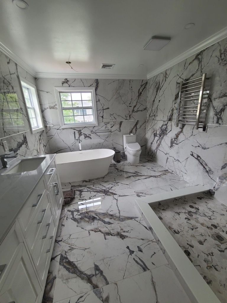 Modern bathroom with white marble tile, a freestanding tub, vanity, toilet, and shower bench.