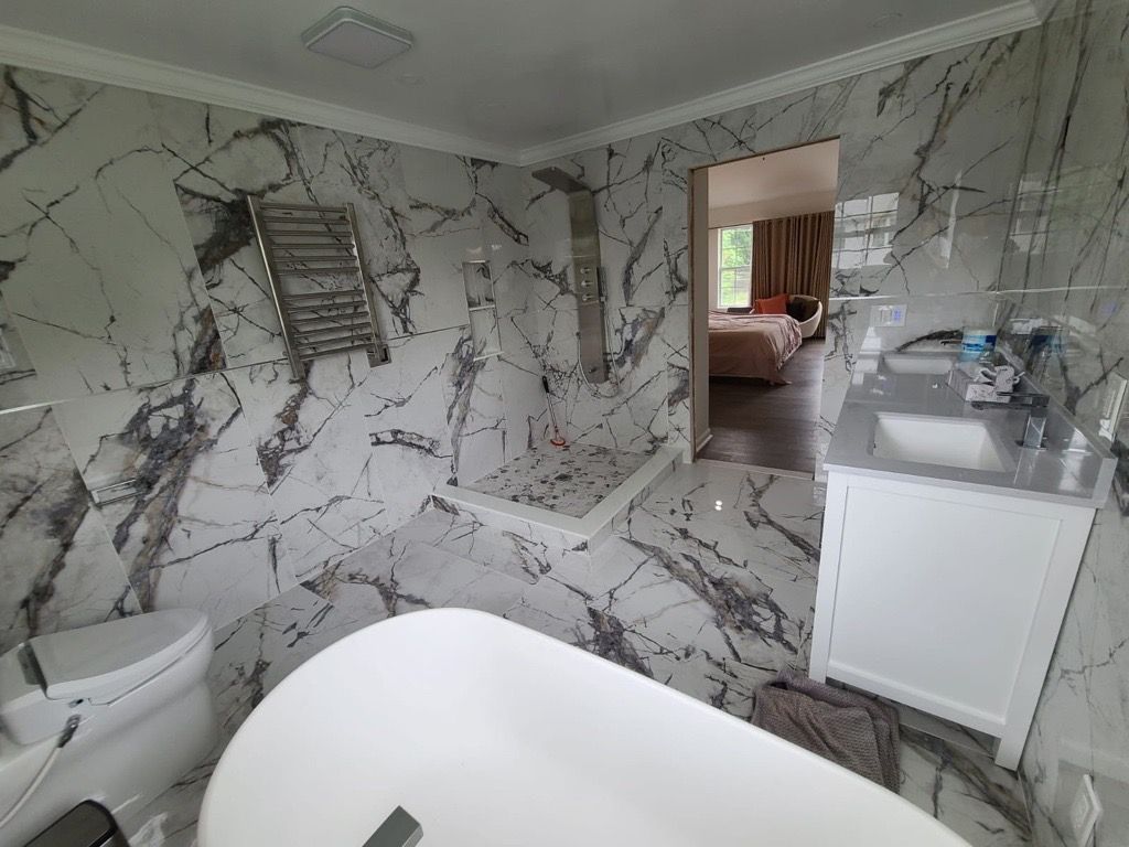 Modern bathroom with marble-pattern walls, white tub, toilet, sink cabinet, and a doorway to a bedroom.