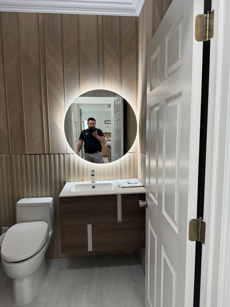 Small bathroom with toilet, wood vanity, lit oval mirror, and a person taking a mirror selfie.