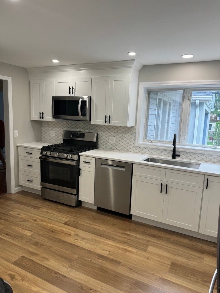 Modern white kitchen with stainless steel appliances, wood floors, and a large window over the sink