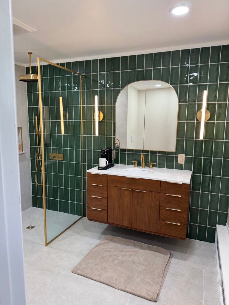 Modern bathroom with green tile wall, wood vanity, round mirror, gold lights, and glass shower divider.
