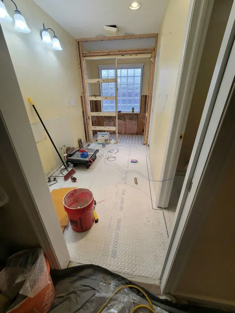 Narrow hallway under renovation with tools, a red bucket, floor covering, and a window at the end.