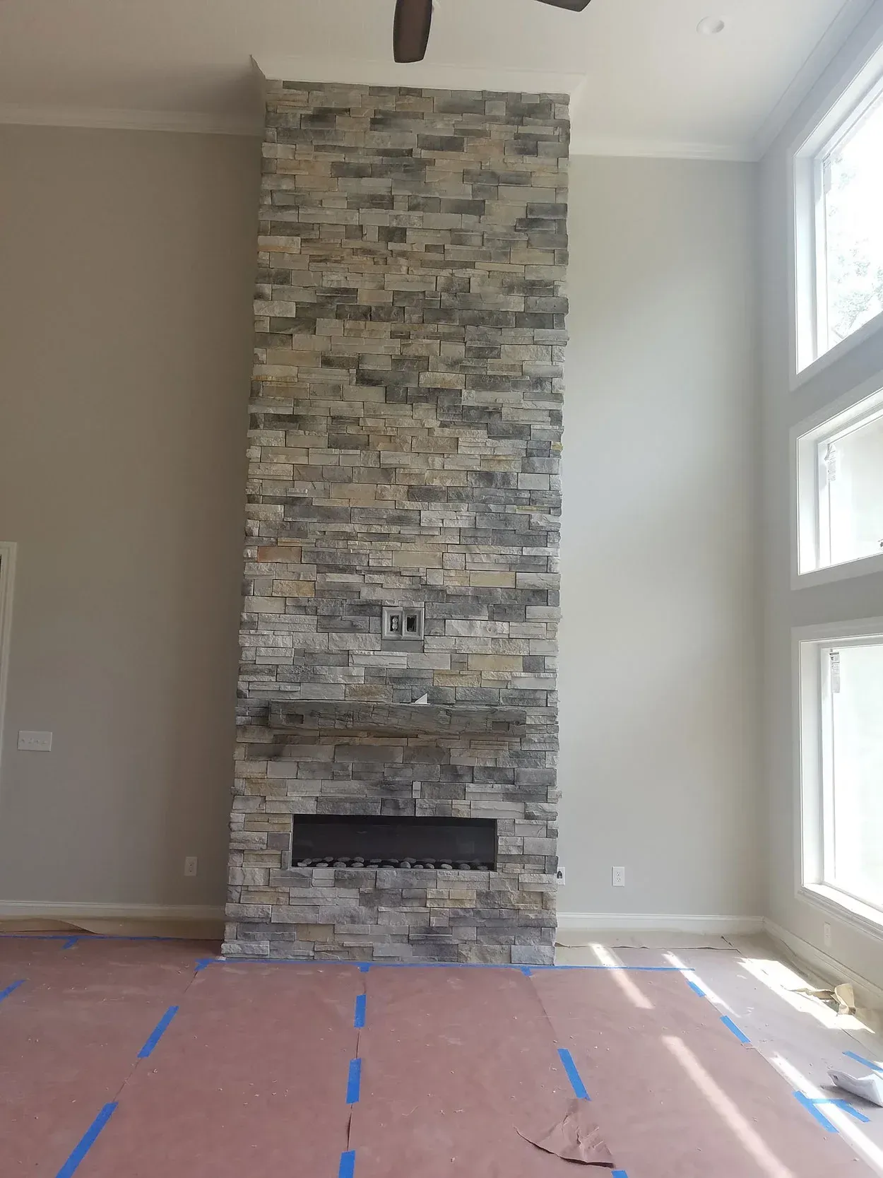 Living room with a tall stacked-stone fireplace and large windows, floor covered with protective tape