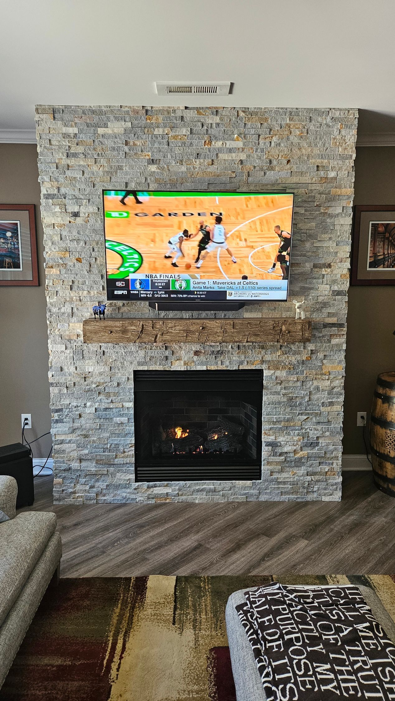 Living room with stone fireplace, mounted TV, and dark floor rug; sports game playing on screen