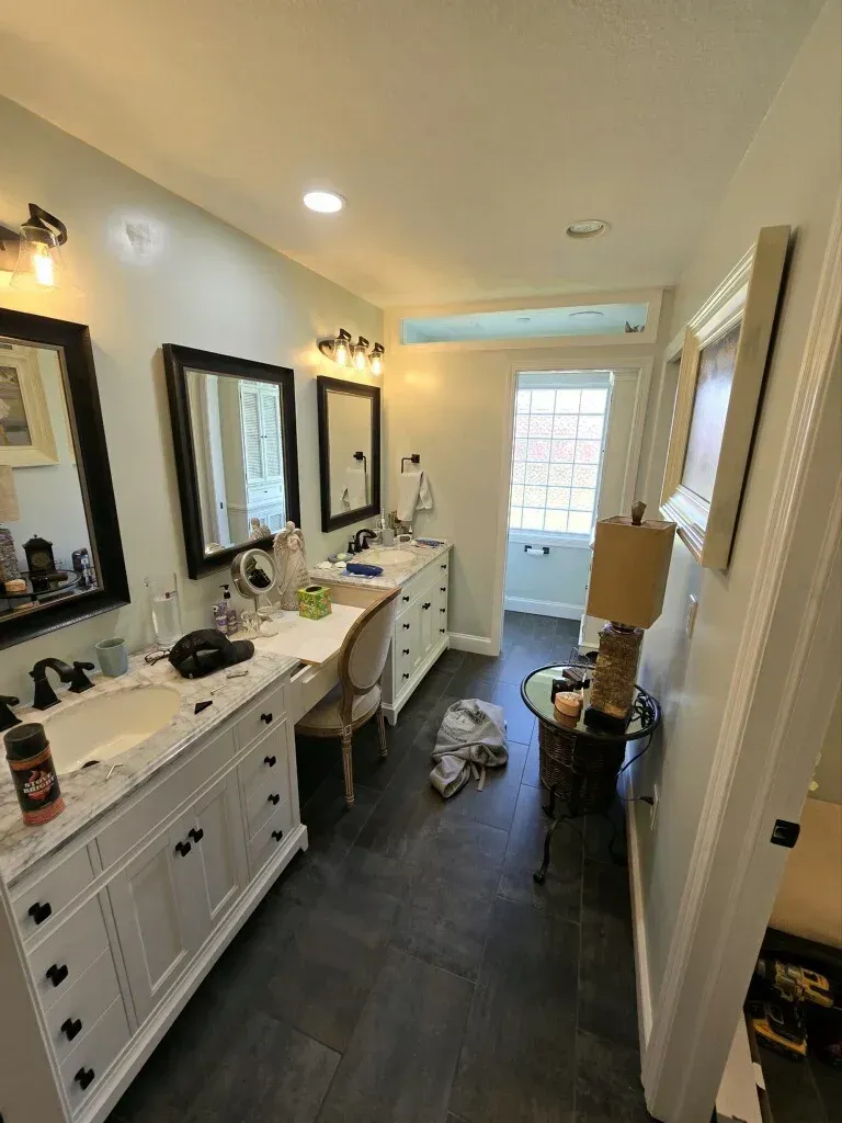 Bright bathroom with double vanity, mirrors, sink clutter, and a glass door at the far end.