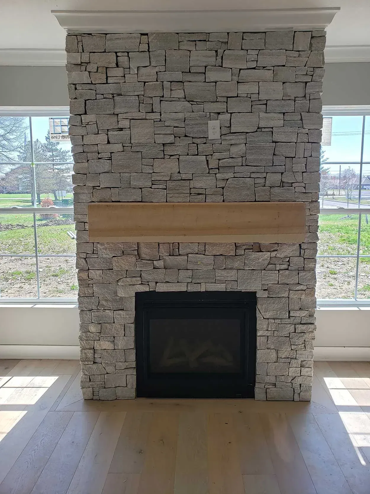 Stone fireplace with a wooden mantel between large windows in a bright room.