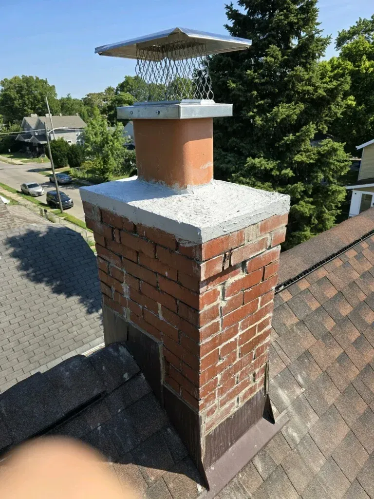 Brick chimney with metal cap on a shingled roof, viewed from above in a sunny neighborhood.