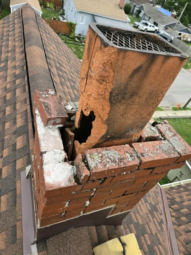 Rusty chimney flue on a brick rooftop, with damaged masonry and a shingled roof in the background.
