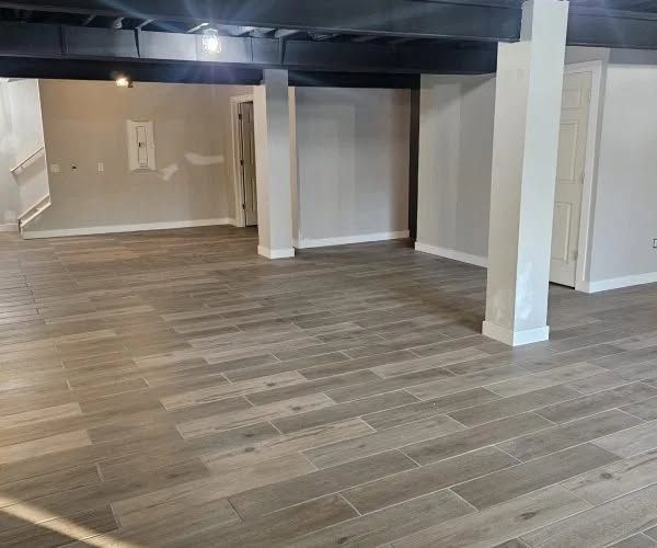 Empty basement room with gray wood-look flooring, white walls, and support columns