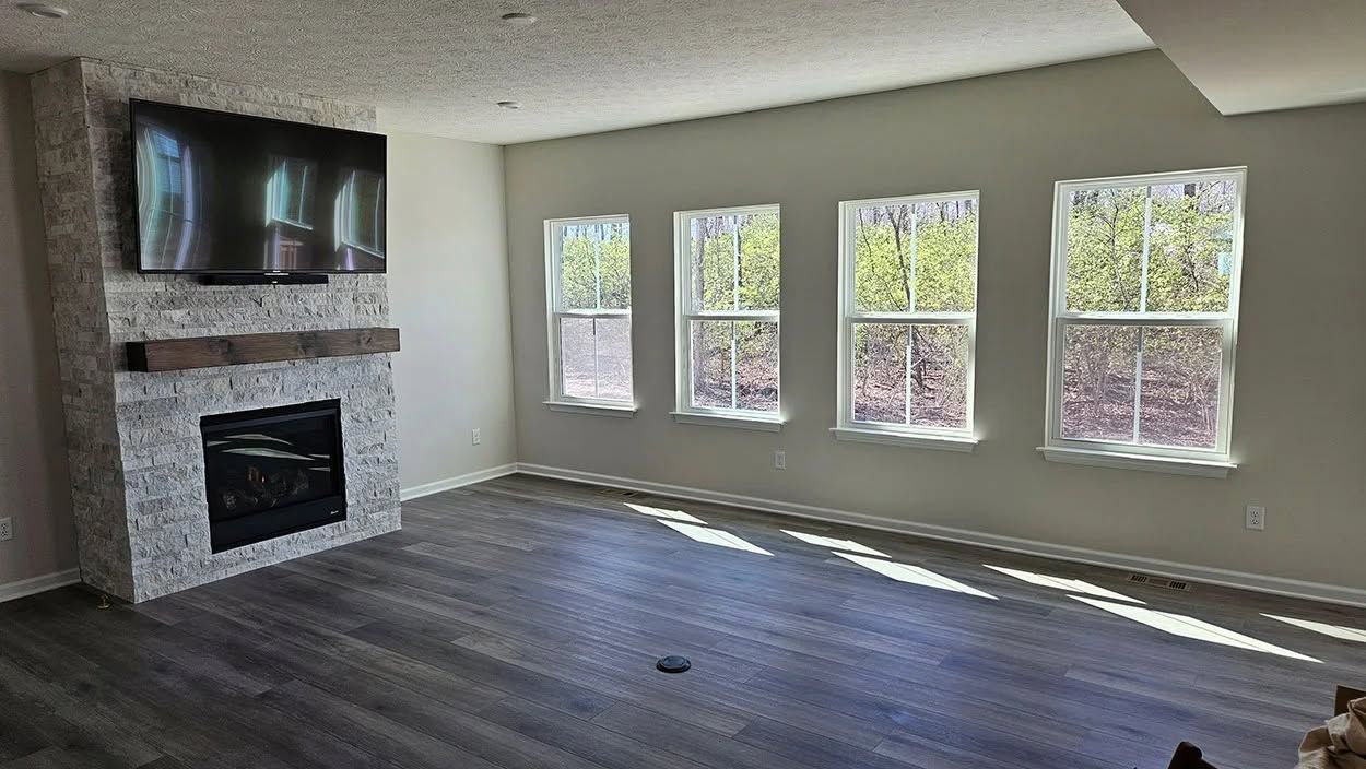 Empty living room with dark wood floors, stone fireplace, and large windows letting in bright daylight