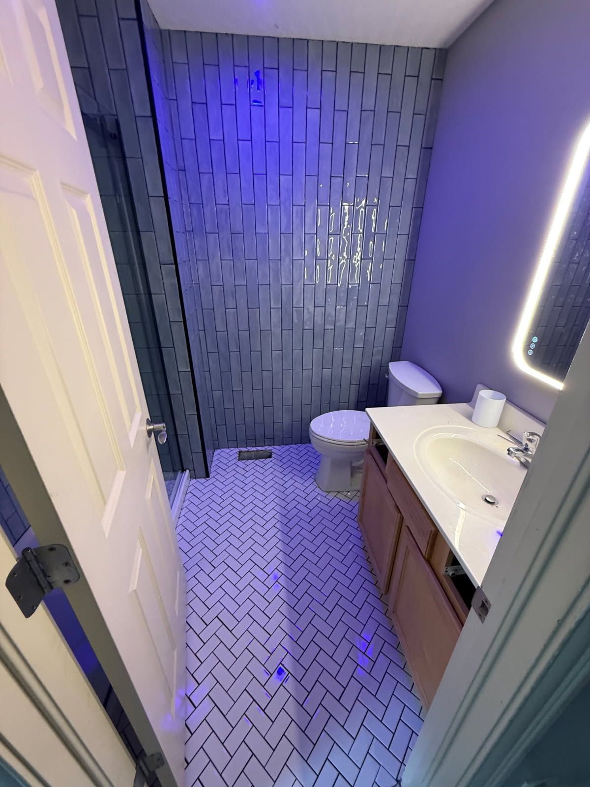 Small bathroom with white vanity, toilet, patterned floor, and blue-lit tiled shower wall
