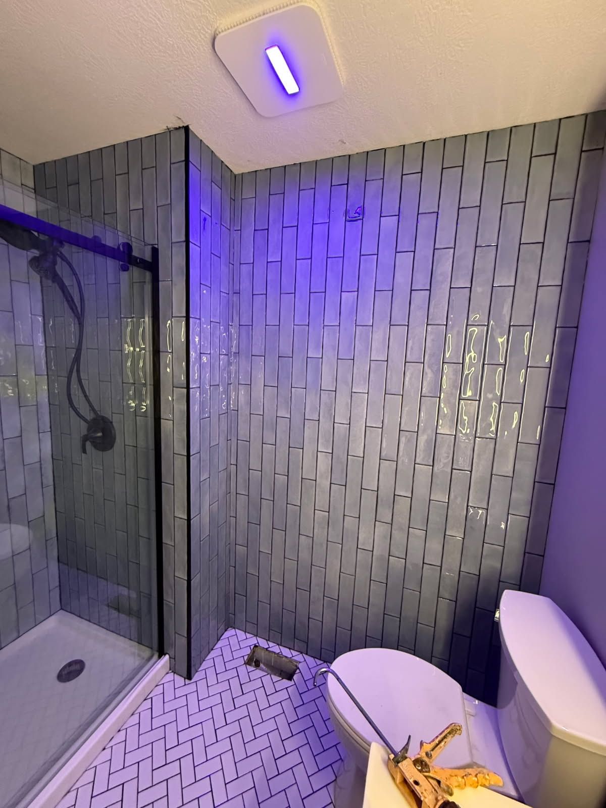 Compact bathroom with glass shower, white tiled walls, and purple lighting.