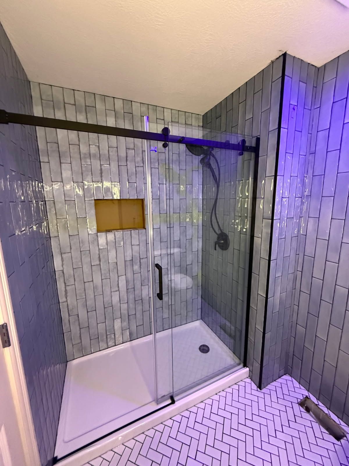 Modern shower with glass doors, gray tile walls, white herringbone floor, and a gold-framed niche.