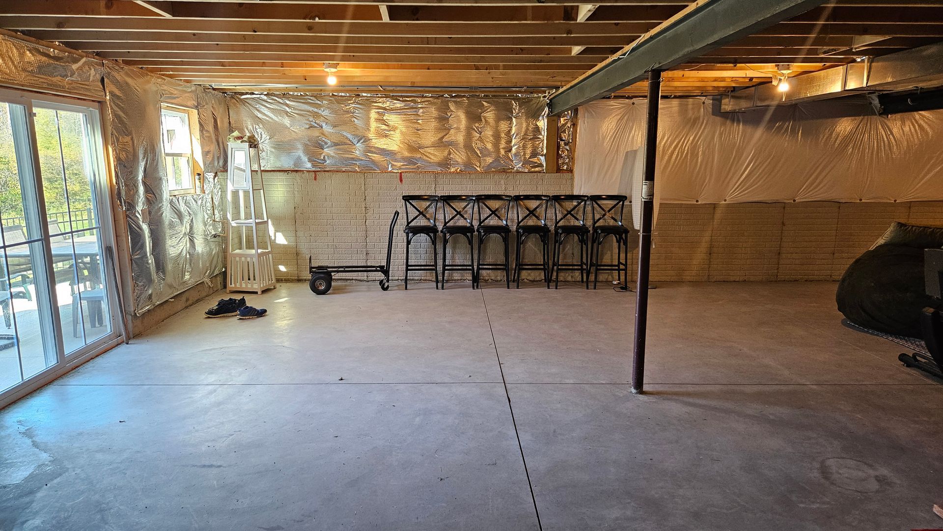 Unfinished basement with exposed beams, concrete floor, plastic-covered walls, and storage shelves along the back wall