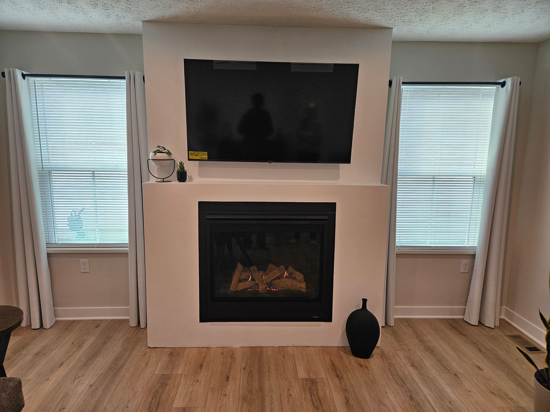 Living room fireplace with wall-mounted TV, two windows with blinds, and a black vase on the floor