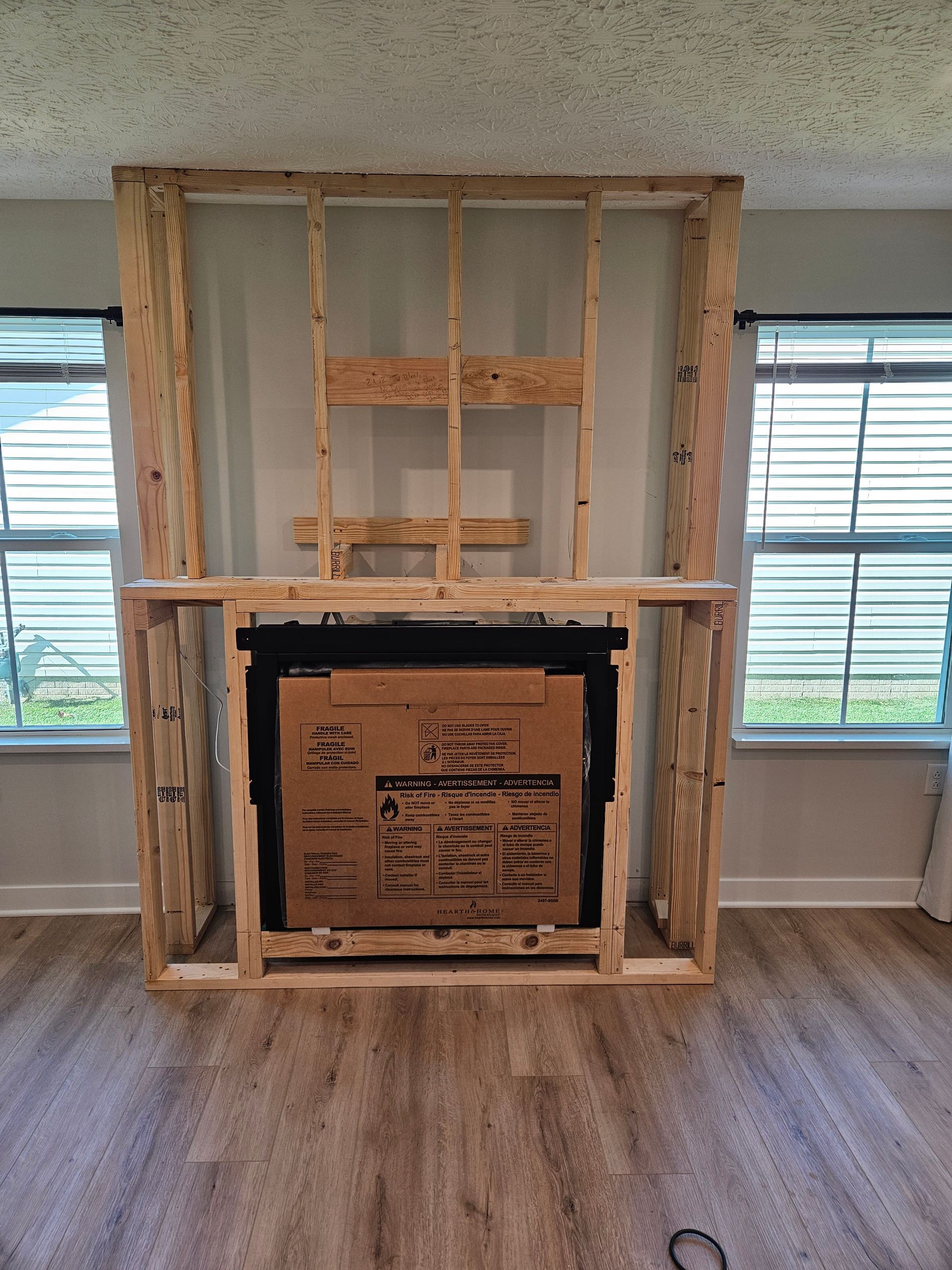 Unfinished wood-framed wall around a fireplace opening with a boxed insert, in a room with windows and hardwood floor