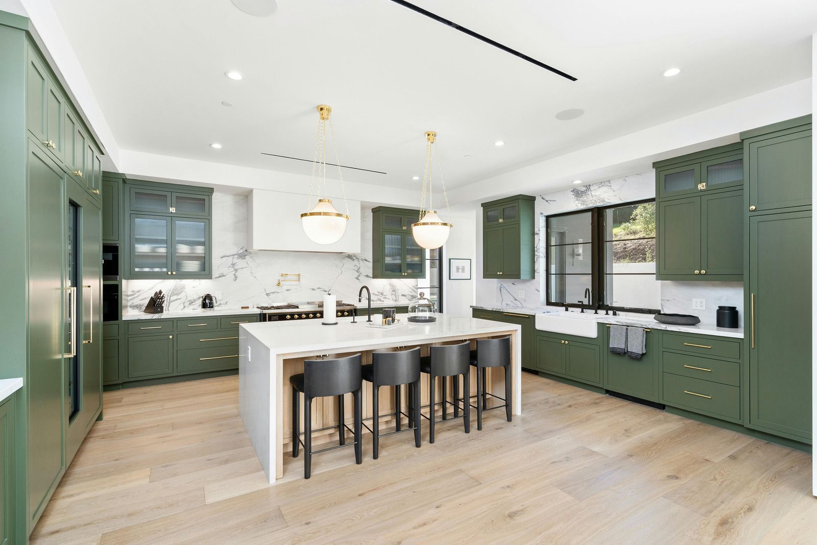 Modern green kitchen with a large island, bar stools, pendant lights, and light wood floors.