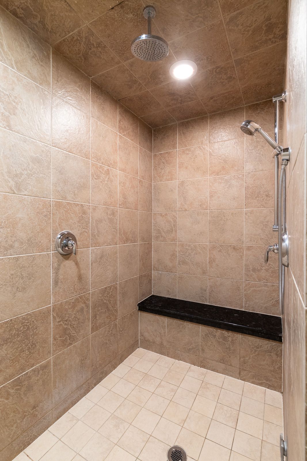 Empty tiled shower with overhead and handheld showerheads, tan walls, white floor, and a black bench.