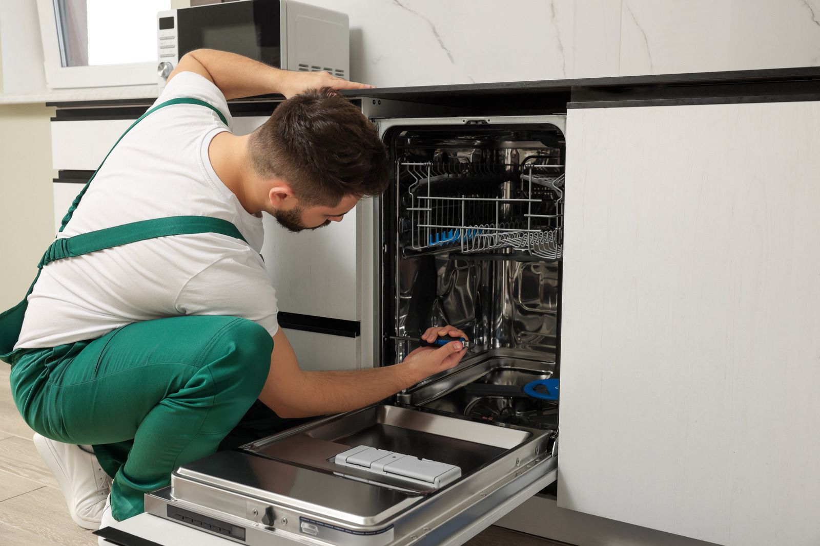 Technician repairing an open dishwasher in a kitchen, crouched and using tools.
