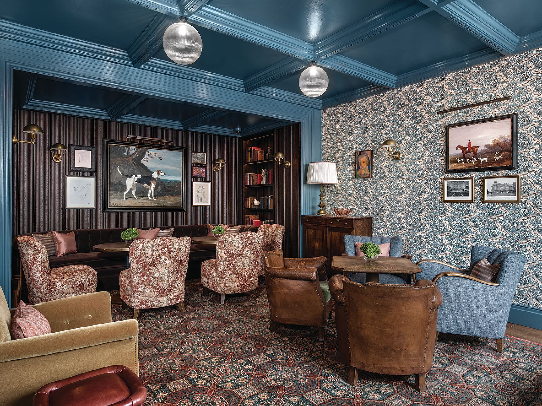 Cozy vintage lounge with patterned armchairs, blue walls, wood paneling, and framed artwork