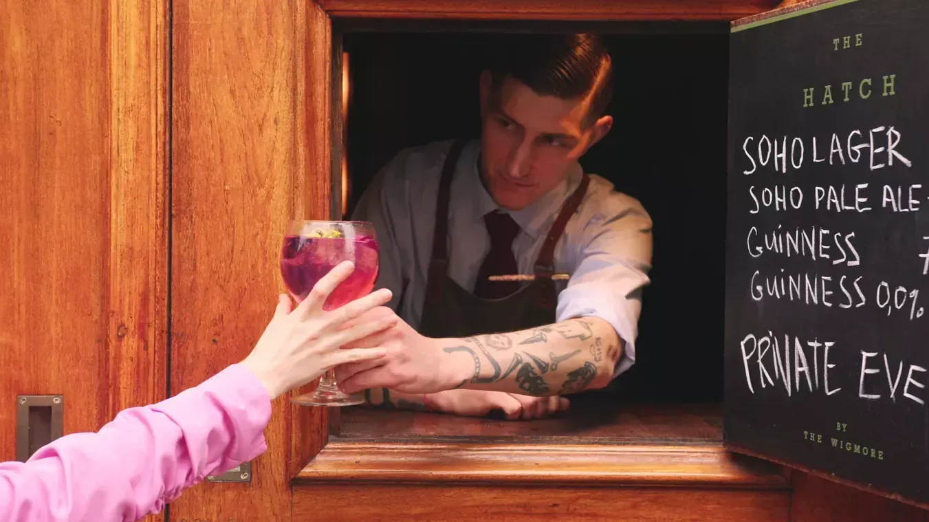 Bartender passing a pink drink through a wooden bar window, with a chalkboard menu beside him