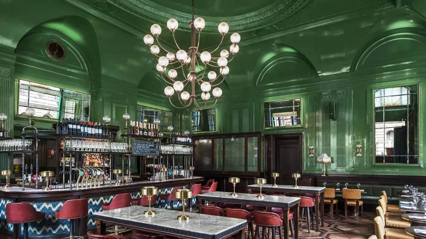Green-paneled elegant bar interior with chandeliers, mirrored walls, and red stools and seating