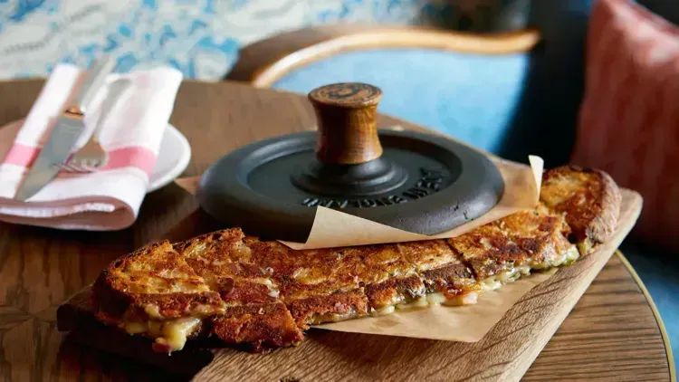 Two slices of cheesy flatbread on a wooden board beside a lidded black pot and folded napkins