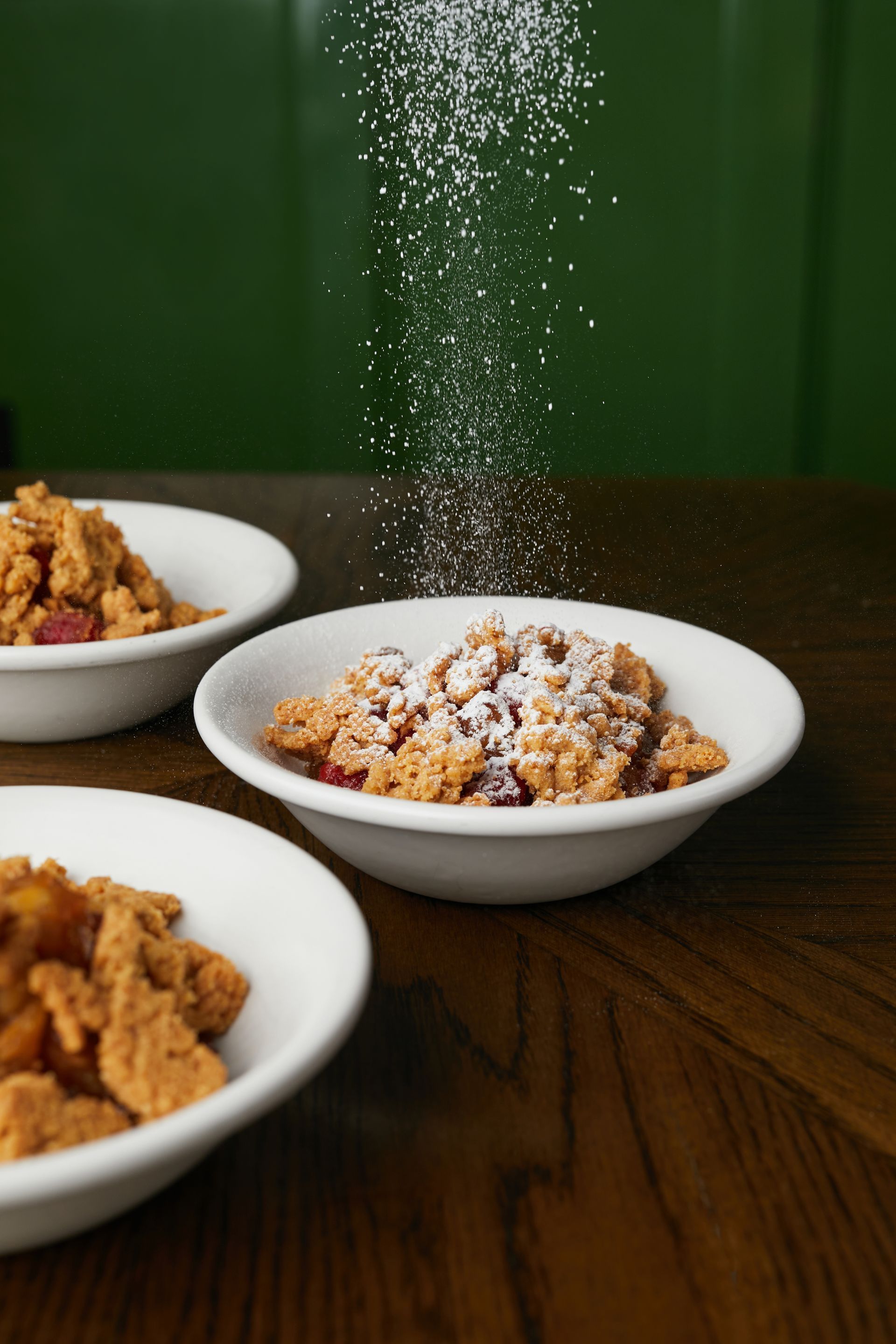 Three bowls of fried chicken on a wooden table, with powdered sugar falling onto the center bowl.