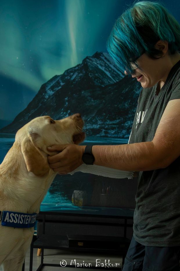Iemand leunt met zijn voorhoofd tegen een grijze, gevlekte hond met open bek, waarmee een moment van stille verbondenheid wordt vastgelegd.