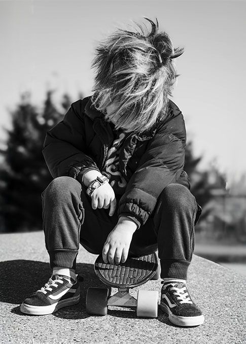 Een persoon met warrig haar zit buiten op een skateboard, met gebogen hoofd, gekleed in een donkere jas en sneakers.