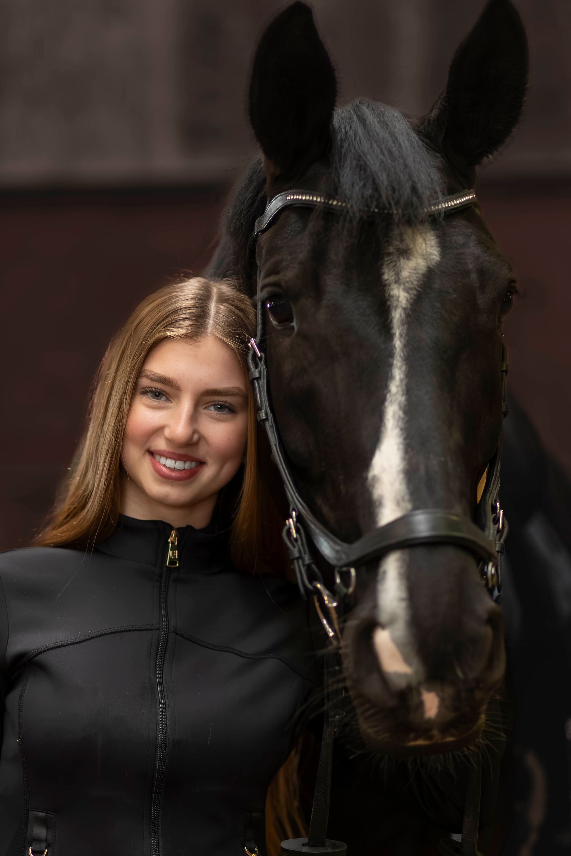 Een lachend persoon met lang haar poseert dicht naast een zwart paard met een hoofdstel.