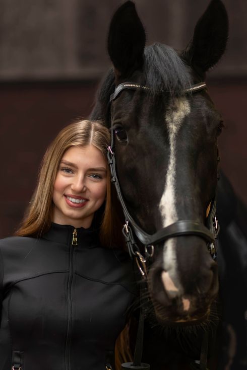 Een lachend persoon met lang haar poseert dicht naast een zwart paard met een hoofdstel.