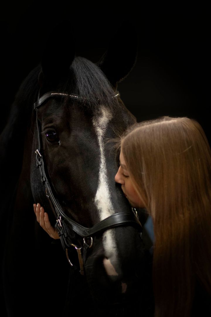 Een persoon leunt zachtjes tegen een donker paard met een witte streep op zijn gezicht, tegen een effen zwarte achtergrond.