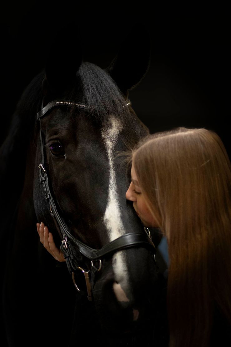 Een persoon leunt zachtjes tegen een donker paard met een witte streep op zijn gezicht, tegen een effen zwarte achtergrond.