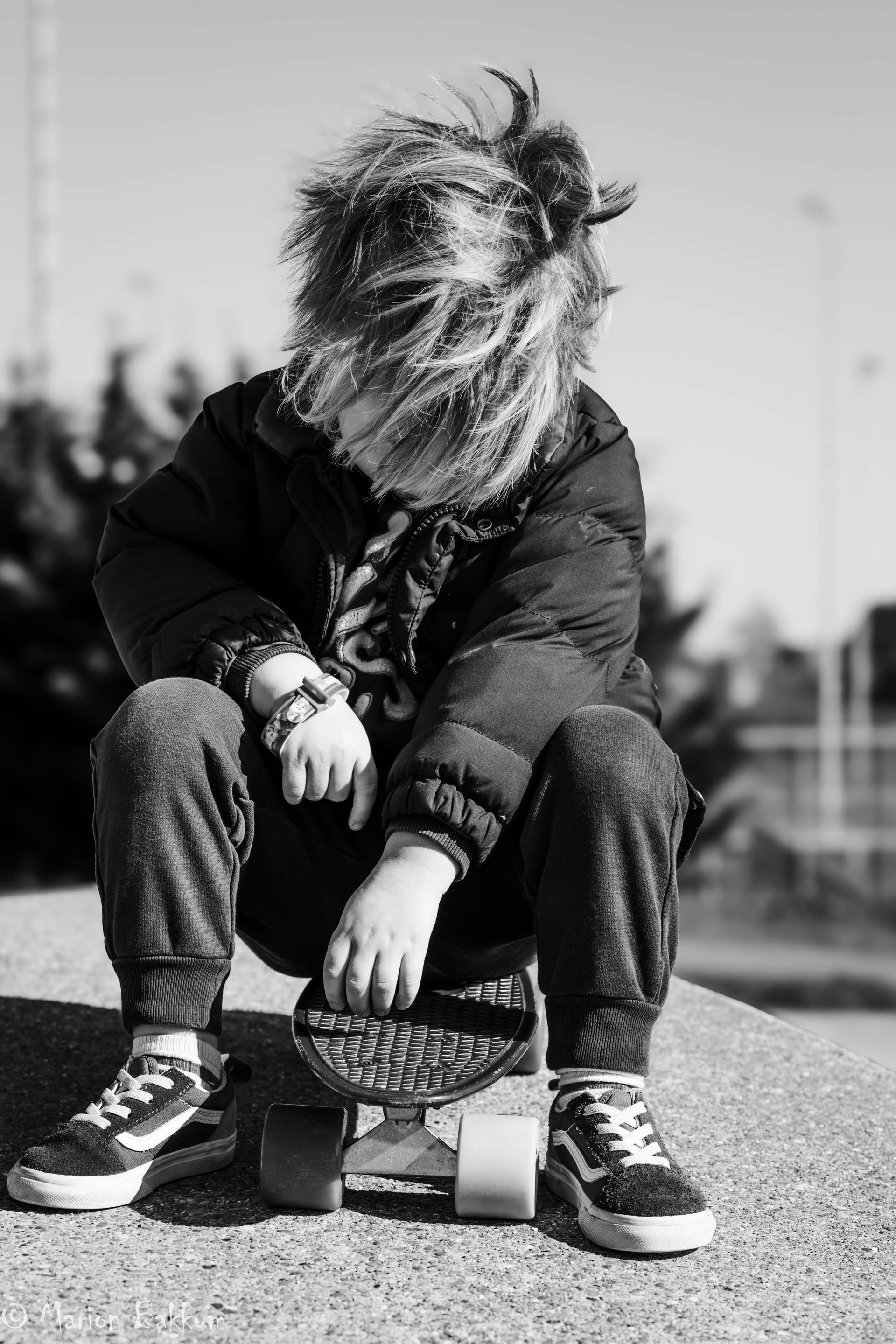 Een persoon met warrig haar zit buiten op een skateboard, met gebogen hoofd, gekleed in een donkere jas en sneakers.