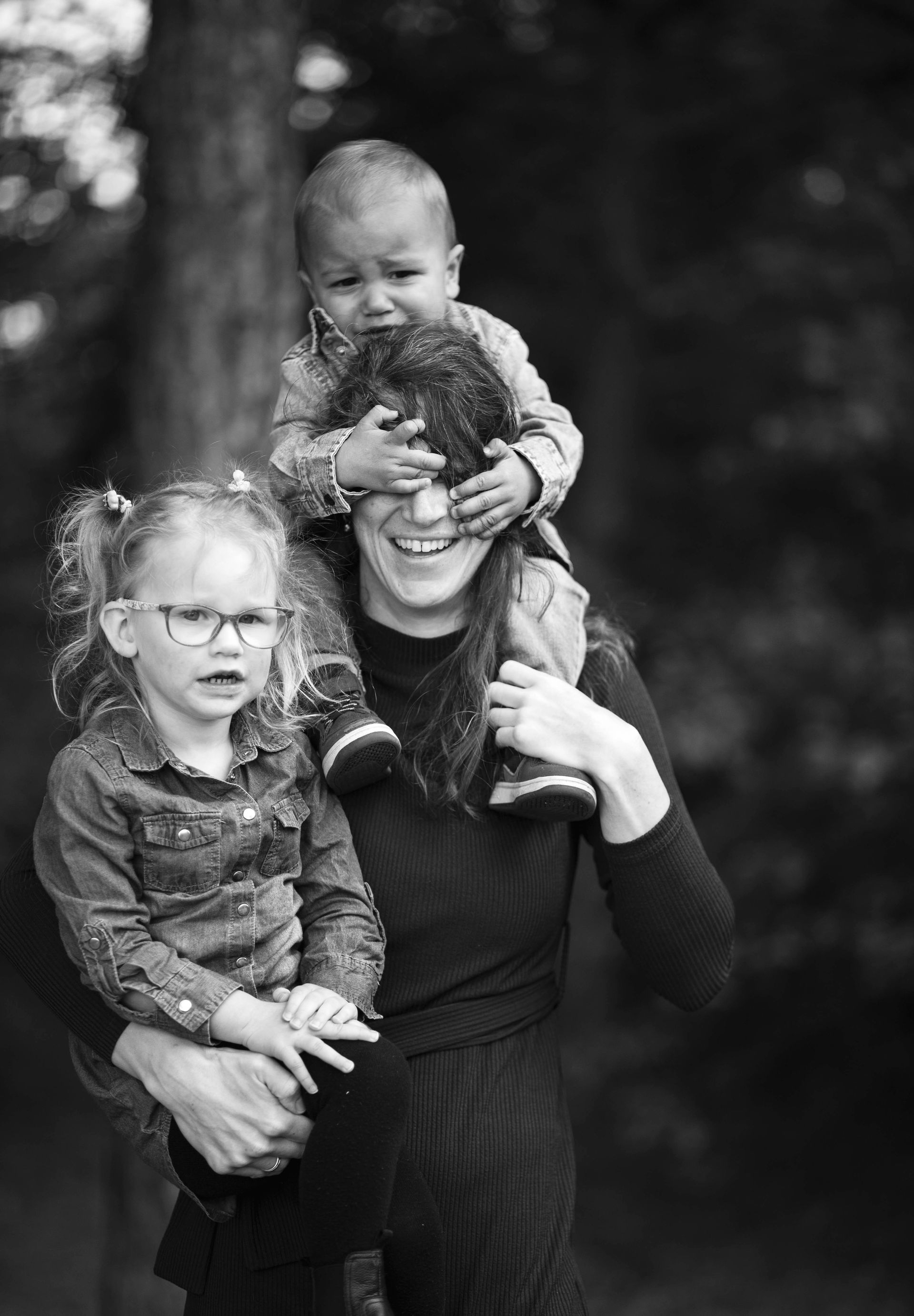 Een persoon draagt een kind op zijn schouders terwijl hij een ander kind vasthoudt, en glimlacht terwijl het kind bovenop zijn ogen bedekt.
