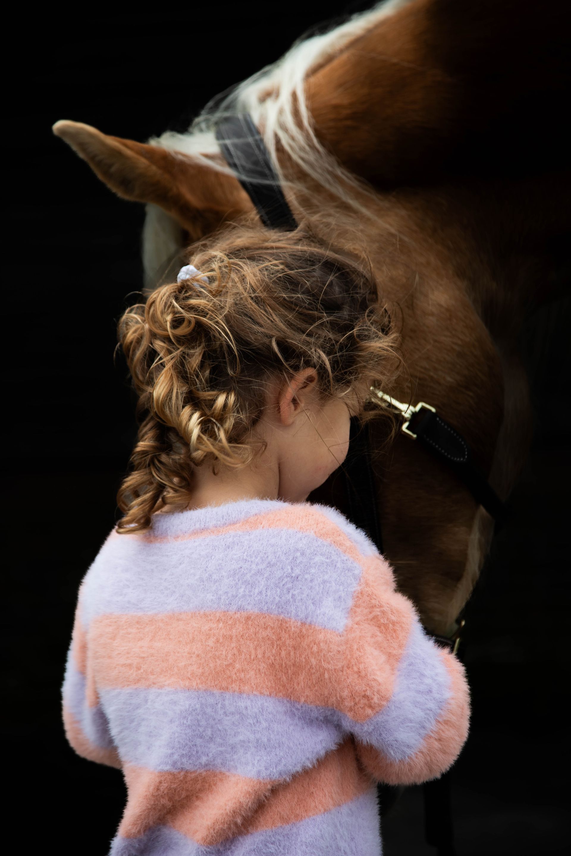 Een kind met krullend haar en een gestreepte trui staat dicht bij een paard; hun voorhoofden raken elkaar tegen een donkere achtergrond.