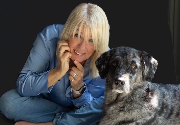 Een persoon in een blauw shirt rust met de kin op de hand naast een gevlekte grijze hond tegen een donkere achtergrond.