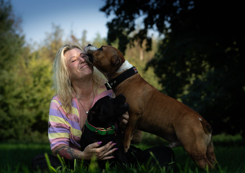 Een persoon in een pastelkleurig gestreept shirt glimlacht terwijl hij of zij wordt gelikt door een bruine hond, terwijl een zwarte hond buiten op zijn of haar schoot zit.