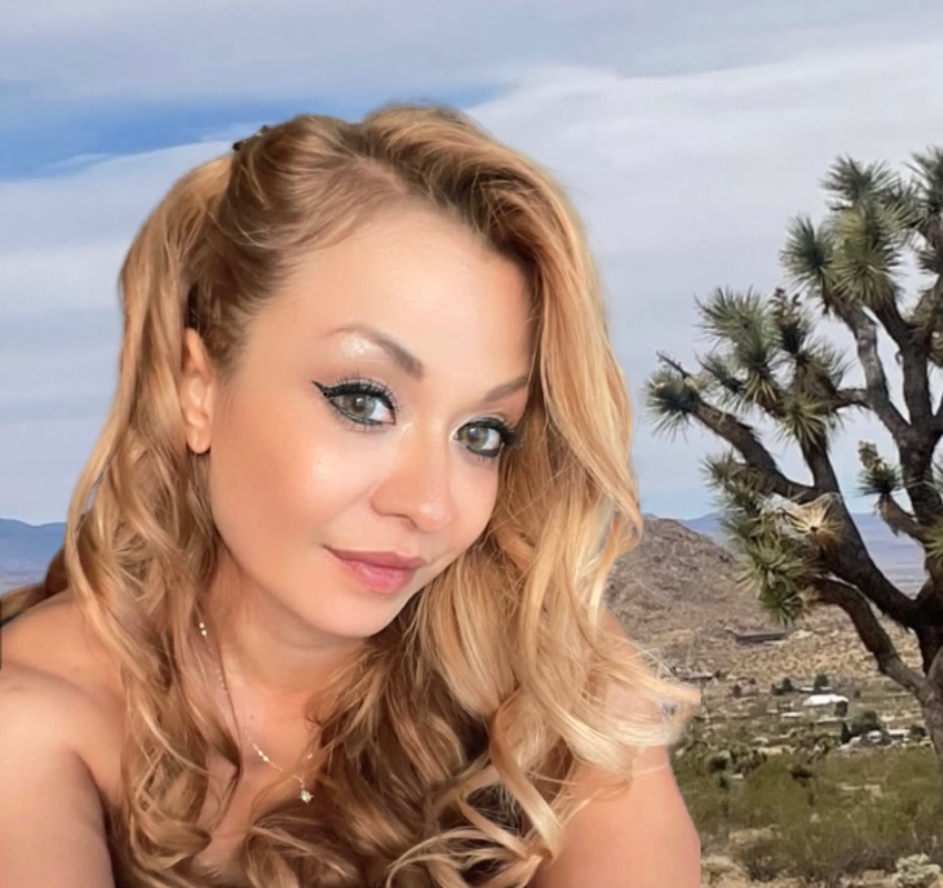 Smiling blonde woman with wavy hair posing outdoors with desert hills and Joshua trees in the background